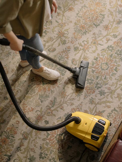 A person operating a yellow and grey vacuum cleaner to deep clean a patterned, beige and light brown upholstered area rug on a wooden floor in a living room. The room features a red leather sofa, and